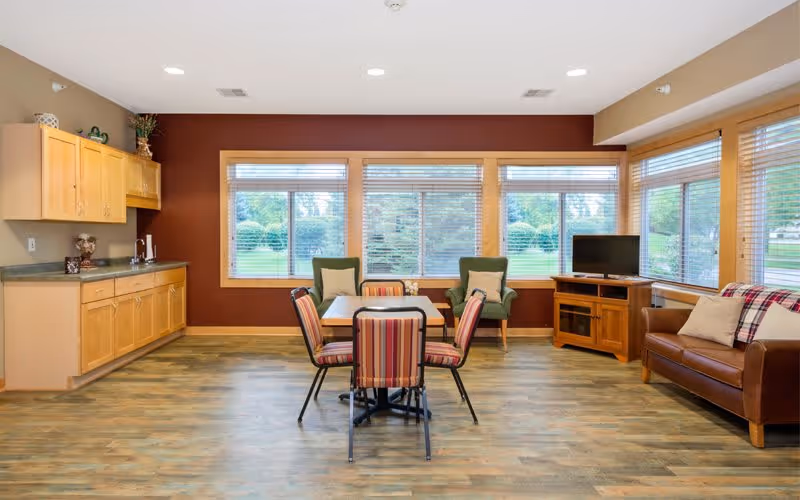 A bright and spacious common area with large windows overlooking greenery. The room features a small dining table with four striped chairs, two green armchairs with pillows, a brown leather sofa with a plaid throw, a wooden TV stand with a flat-screen TV, and a kitchenette with wooden cabinets and a countertop on the left side.