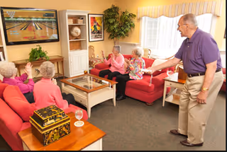 A group of elderly residents in a cozy living room playing a video bowling game on TV while seated on red sofas.