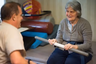 An elderly woman holding a gray exercise bar while sitting on a padded surface, engaging in a physical therapy or exercise session with a man who is facing her.