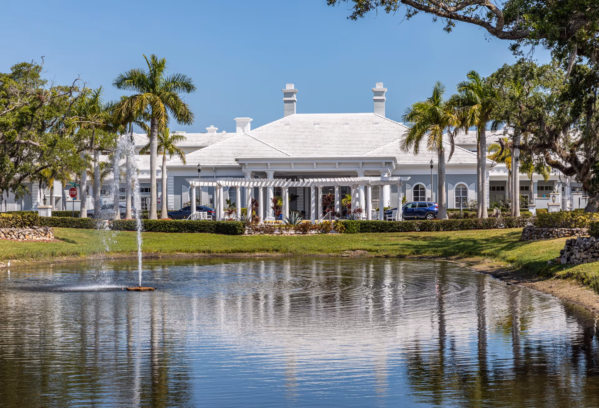 View of a senior living facility building with a white roof and columns, surrounded by palm trees and greenery, with a pond and a water fountain in the foreground under a clear blue sky.