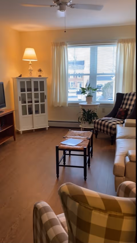 A cozy living room with wooden flooring, a white cabinet with glass doors and a lit lamp on top, a window with cream curtains letting in natural light, a small round table with potted plants, a checkered armchair, a beige sofa, and two small stools in the center.