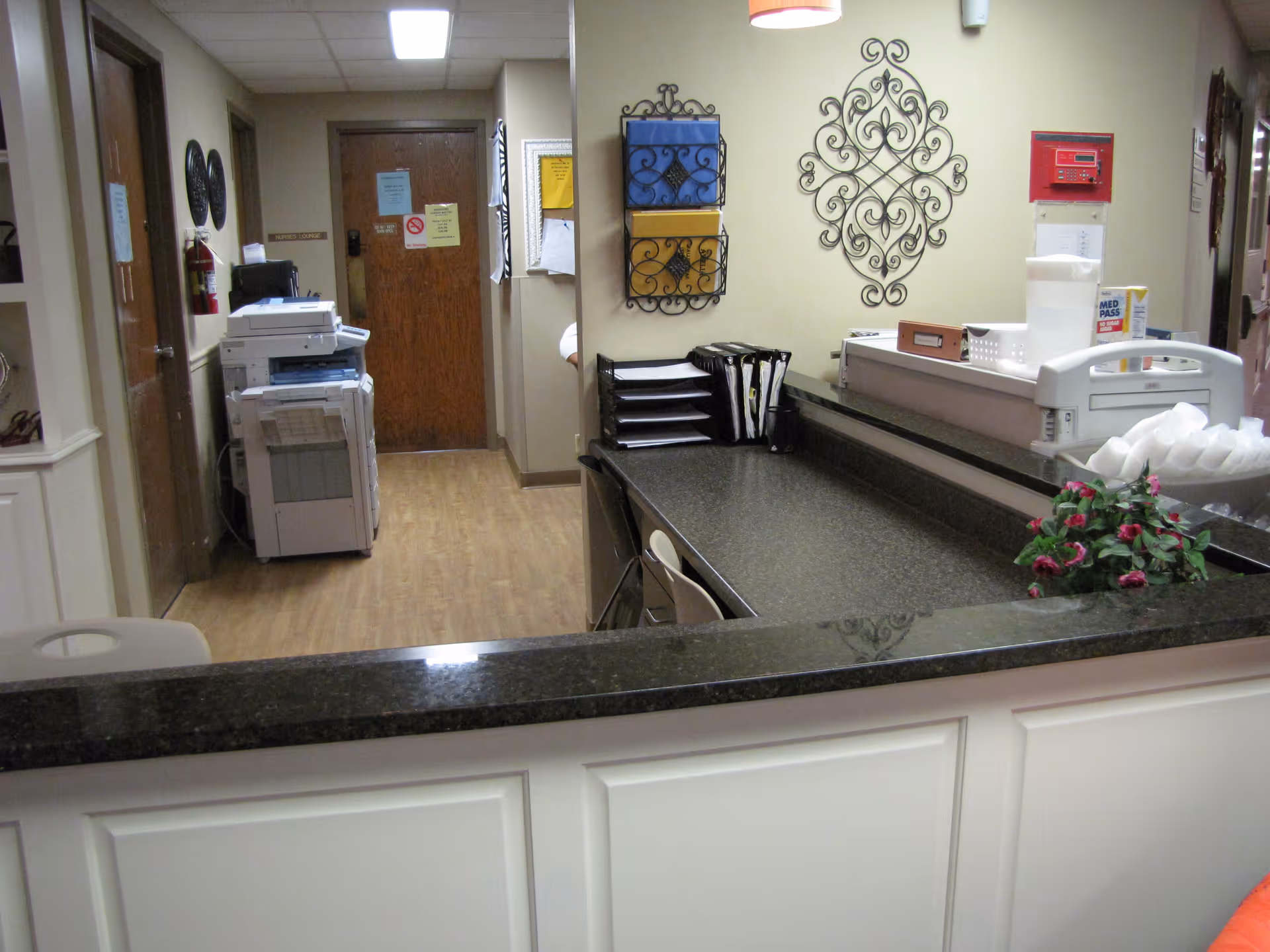 Reception desk area with a black countertop and white paneling, a chair tucked under the desk, wall-mounted decorative metal art, a printer/copier machine in the hallway, and various office supplies including a tissue box and file organizers.