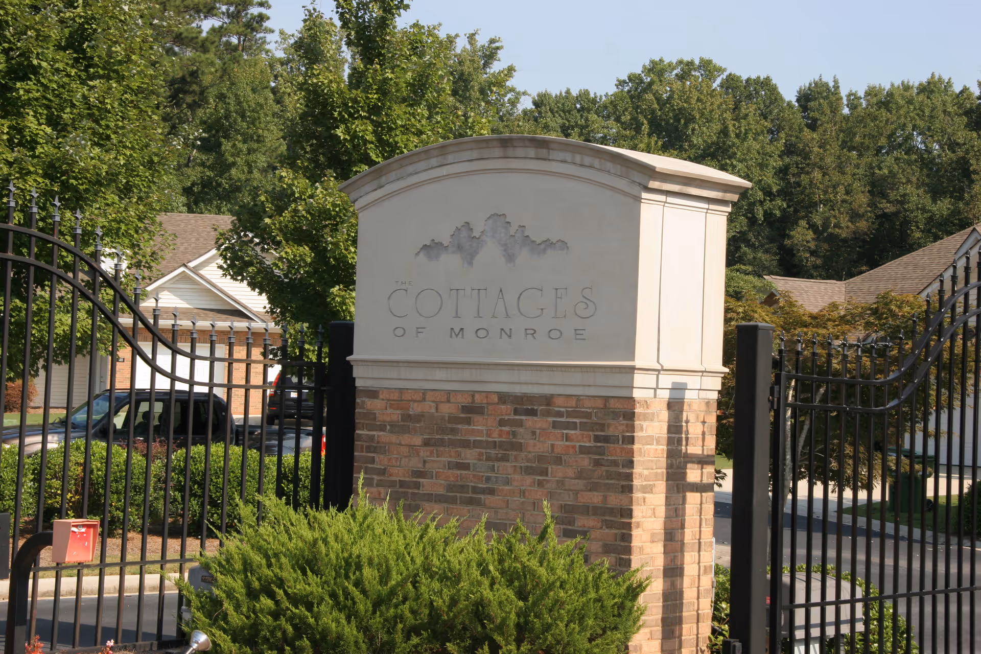 Entrance sign for The Cottages of Monroe, a senior living facility, with a brick and stone pillar surrounded by greenery and black metal gates. Residential buildings and trees are visible in the background.