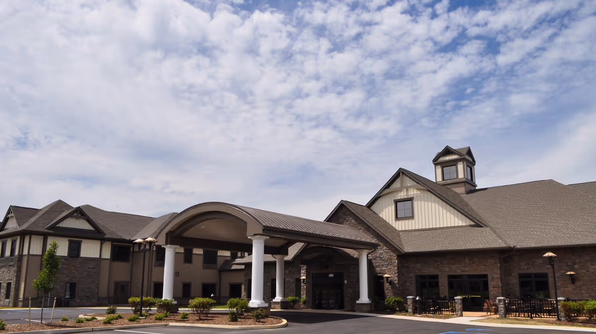 Front entrance of a large retirement community building with a covered porte-cochère, white columns, stone facade and landscaped driveway under a partly cloudy sky.