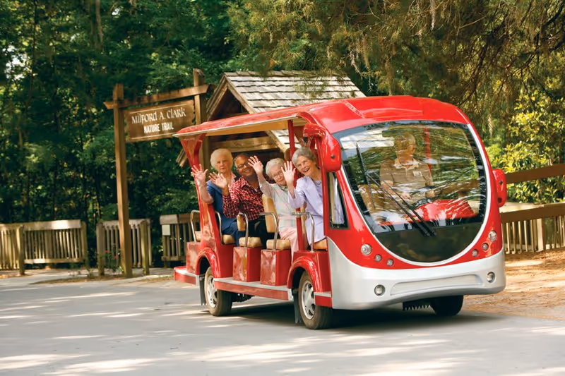 A group of elderly people riding a small red and white open-air electric tram on a paved path near a wooden sign that reads 'Milford A. Clark Nature Trail' surrounded by trees and greenery.