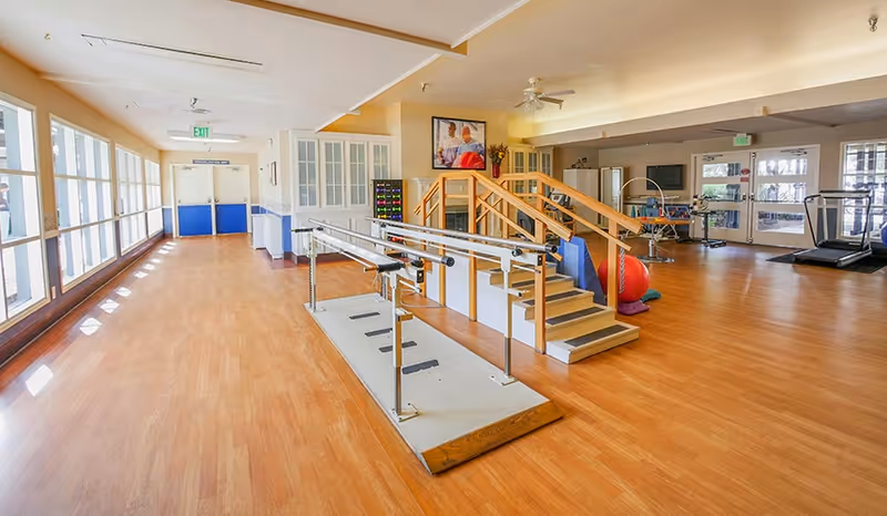 A spacious indoor rehabilitation gym with parallel bars, practice stairs, exercise balls, and treadmills.