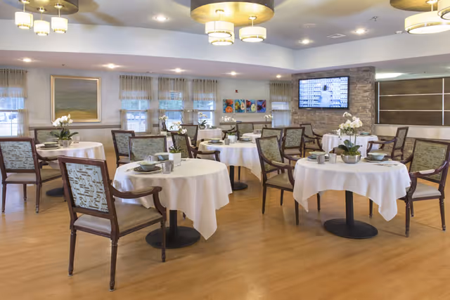 A dining room with round tables covered in white tablecloths, each set with plates, bowls, and cups. Wooden chairs with patterned upholstery surround the tables. The room has wooden flooring, large windows with sheer curtains, modern ceiling lights, and a wall-mounted television.