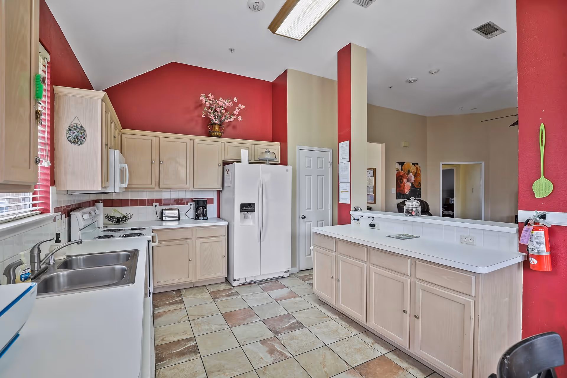 A kitchen area with beige cabinets, a white refrigerator, microwave, stove, and double sink. The walls are painted red and beige, with a vase of flowers on top of the cabinets. The floor is tiled with a mix of beige and brown tiles. There is a fire extinguisher mounted on the right wall and a counter with white countertop extending into the room.