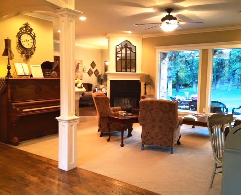 A cozy living room with two patterned armchairs facing a fireplace with a decorative mirror above it. To the left, there is a wooden piano with a clock and lamp on top. Large windows on the right side show a green outdoor area with trees and patio furniture. The room has a warm, inviting atmosphere with soft lighting and a mix of carpet and hardwood flooring.