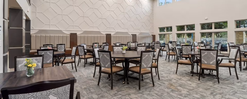 Spacious, well-lit dining room with round and square tables, upholstered chairs, decorative wall panels, and large windows.
