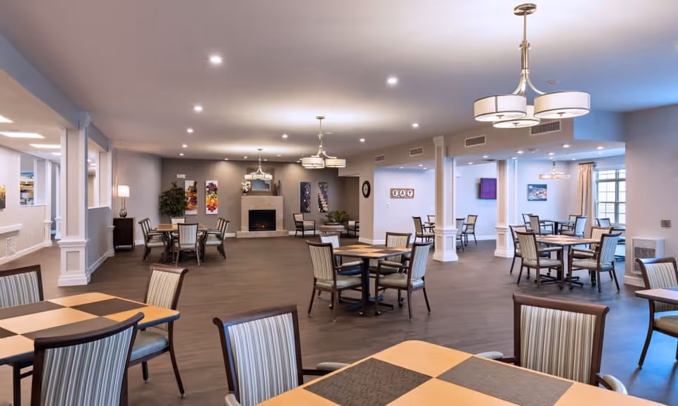 A spacious and well-lit dining area with multiple tables and chairs arranged neatly. The room features a modern fireplace on the far wall, decorative artwork, and several ceiling light fixtures. Large windows allow natural light to enter, and the overall decor is warm and inviting.