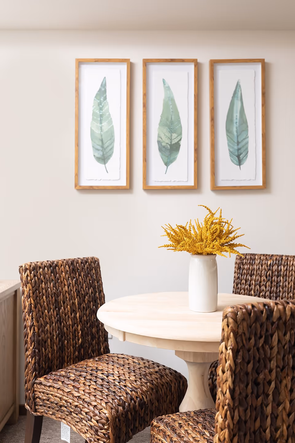A round wooden dining table with a white vase holding yellow flowers in the center, surrounded by four woven rattan chairs. On the wall behind the table are three framed botanical prints of green leaves.