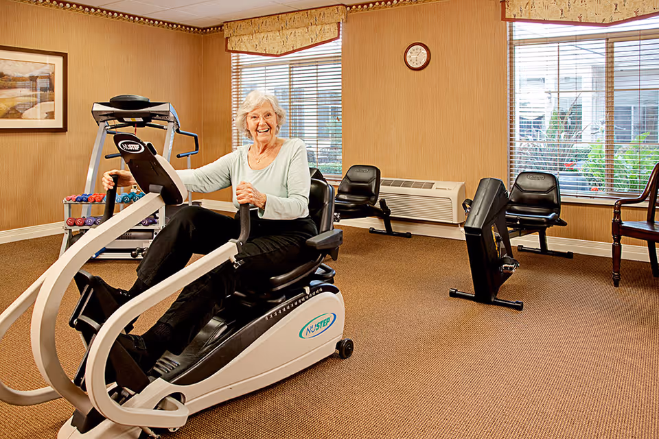 An elderly woman exercising on a NuStep recumbent cross trainer in a well-lit fitness room with beige walls and carpet. The room has large windows with blinds, a clock on the wall, a rack of colorful dumbbells, and additional exercise equipment including rowing machines and chairs.
