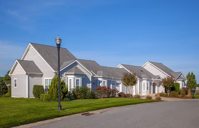 Exterior view of a single-story residential building with light gray siding, multiple gabled roofs, and a well-maintained lawn with shrubs and small trees under a clear blue sky.