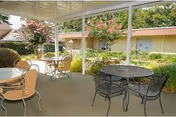 Covered outdoor patio area with several round metal tables and chairs, surrounded by greenery and flowering trees, adjacent to a beige building with windows and doors.