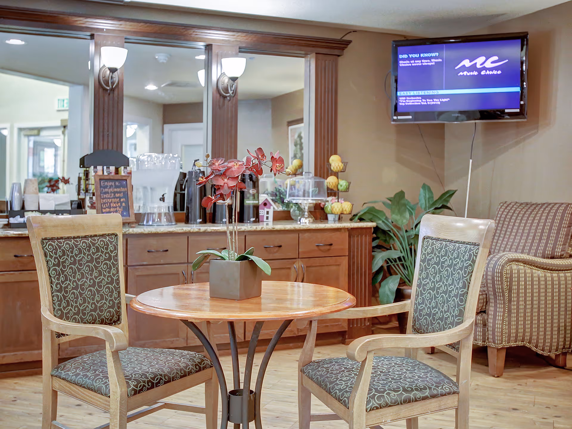 A cozy seating area in a senior living facility with two wooden chairs featuring patterned cushions around a small round wooden table with a potted orchid. Behind the table is a wooden cabinet with a granite countertop holding beverage dispensers, cups, and a small chalkboard sign. A large mirror with wooden trim and wall sconces is mounted above the cabinet. A flat-screen TV is mounted on the wall to the right, and a striped armchair and green plant are visible in the corner.