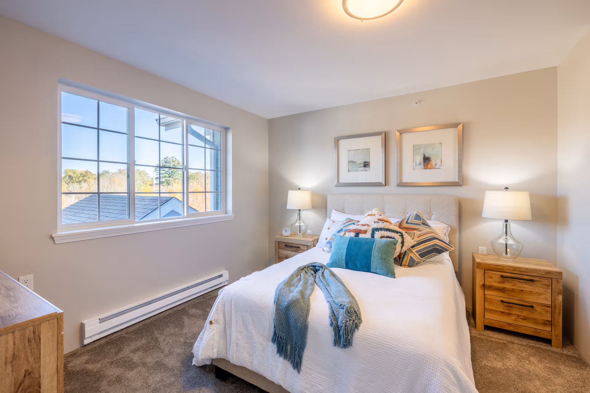 A bright and cozy bedroom with a large window showing an outdoor view. The room features a bed with white bedding, multiple decorative pillows, and a blue throw blanket. There are two wooden nightstands on either side of the bed, each with a lamp. Two framed abstract artworks hang above the bed.