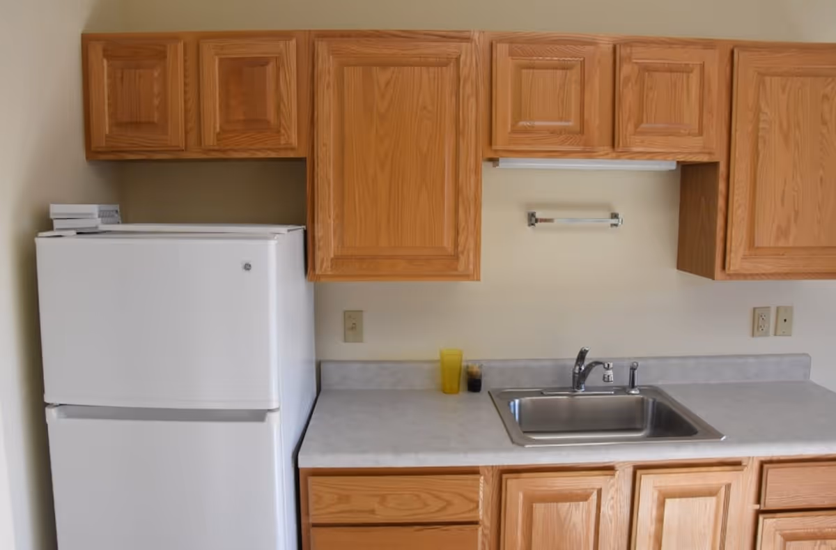 A kitchen area with wooden cabinets, a white refrigerator, a stainless steel sink, and a countertop with a yellow cup and a small dark container.