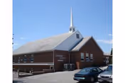 Exterior view of a brick building with a white steeple against a clear blue sky. Several windows are visible along the side, and cars are parked in front of the building.
