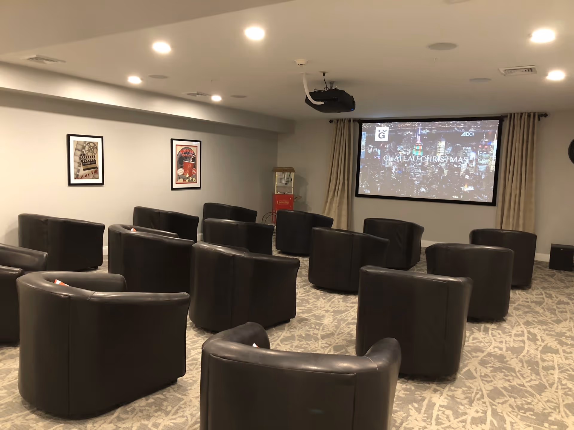 A small theater room with multiple black leather chairs arranged in rows facing a large projector screen displaying a movie titled 'Chateau Christmas'. The room has framed movie posters on the wall and a popcorn machine in the corner.