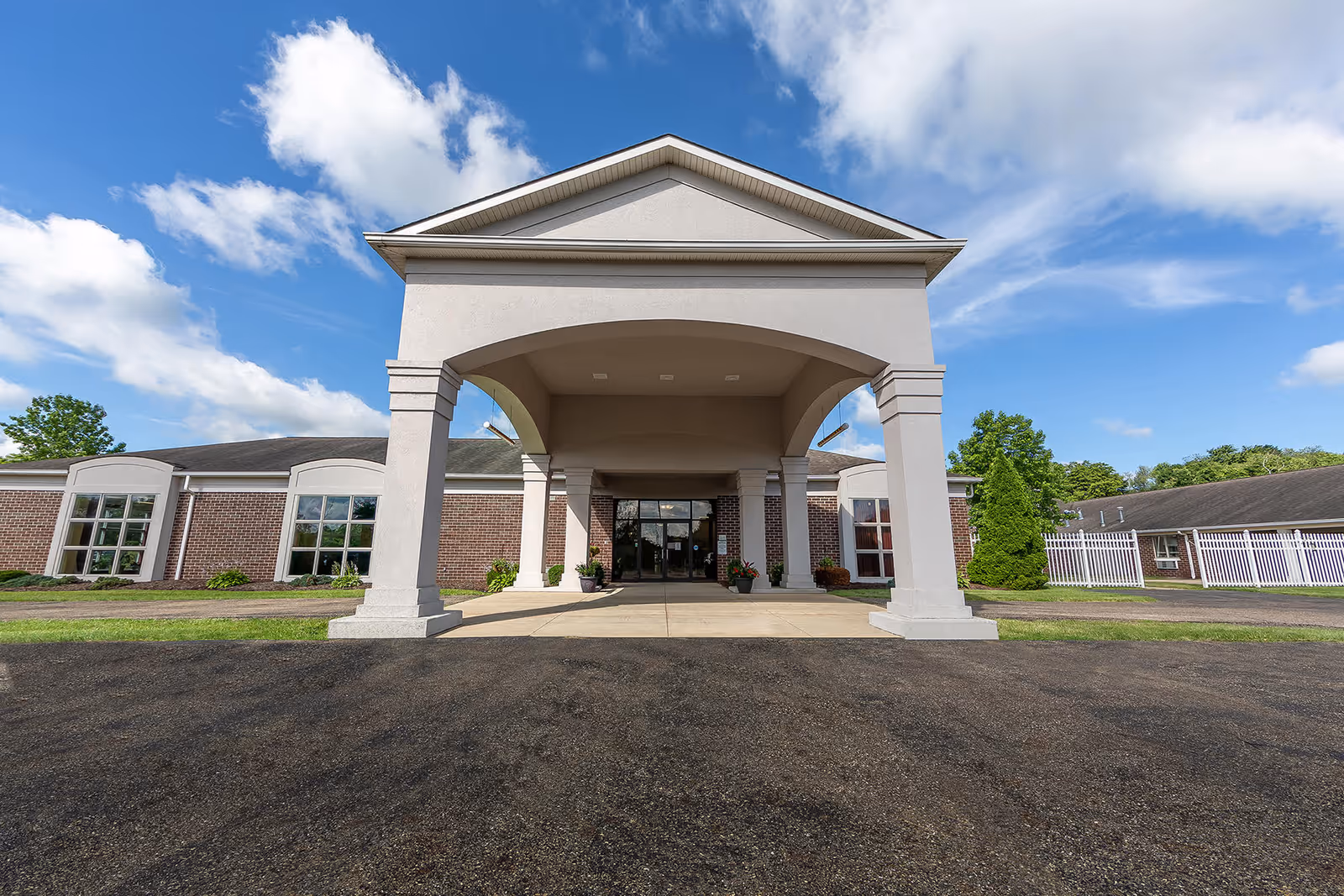 Front exterior view of Lisbon skilled nursing facility showing a large covered entrance with columns, brick walls, multiple windows, and a clear blue sky with some clouds.