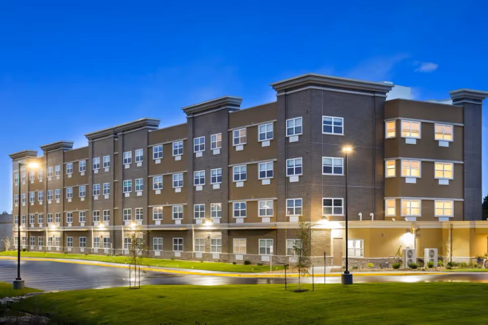 Exterior view of a modern four-story senior living facility building at dusk with lights on inside and street lamps illuminating the parking area and surrounding lawn.