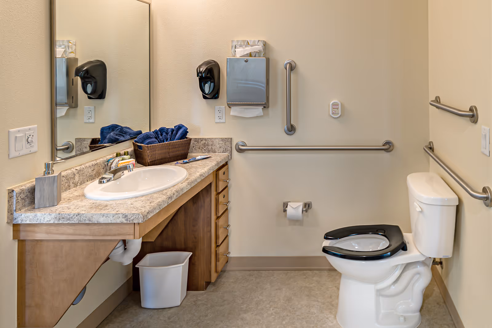 A clean and accessible bathroom with a toilet featuring a raised black seat, grab bars on the walls, a countertop with a sink, a soap dispenser, a tissue dispenser, a basket with blue towels, and a large mirror above the sink.