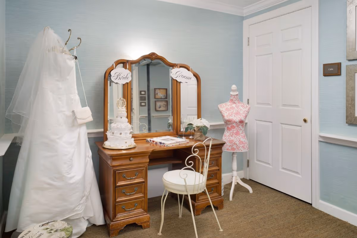 A room with a wooden vanity table and mirror labeled Bride and Groom, a white cushioned chair, a white wedding dress with veil hanging on a stand, a decorative wedding cake on the vanity, a floral mannequin, and a closed white door against light blue walls.