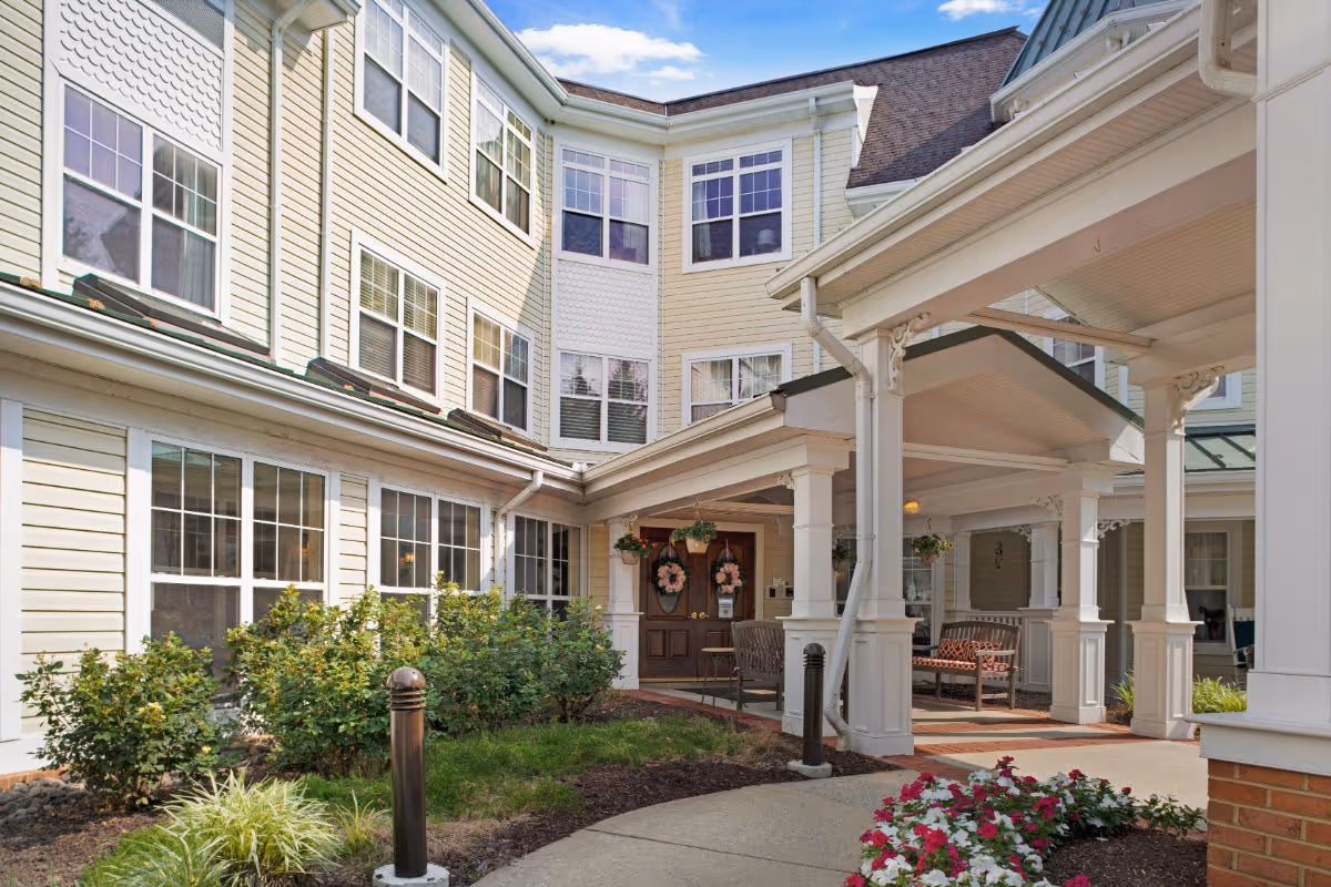 Covered entrance and porte-cochère of a multi-story senior living facility with seating and landscaped flower beds.