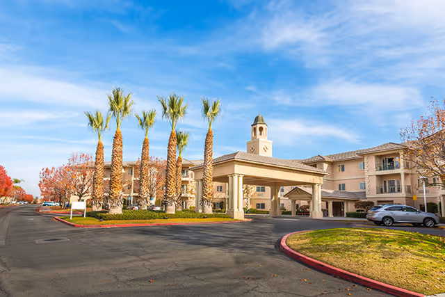 Exterior view of the Riverwalk senior living facility showing a three-story building with a covered entrance, palm trees, and a clear blue sky.