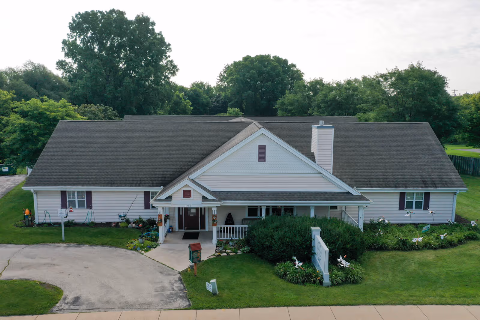 Front exterior view of a single-story assisted living facility building with a gable roof, white siding, and a covered entrance. The building is surrounded by green grass, bushes, and trees in the background.