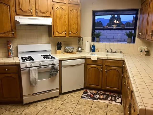 A kitchen with oak cabinets, a white gas stove and dishwasher, tiled countertops and floor, and a window with potted plants.