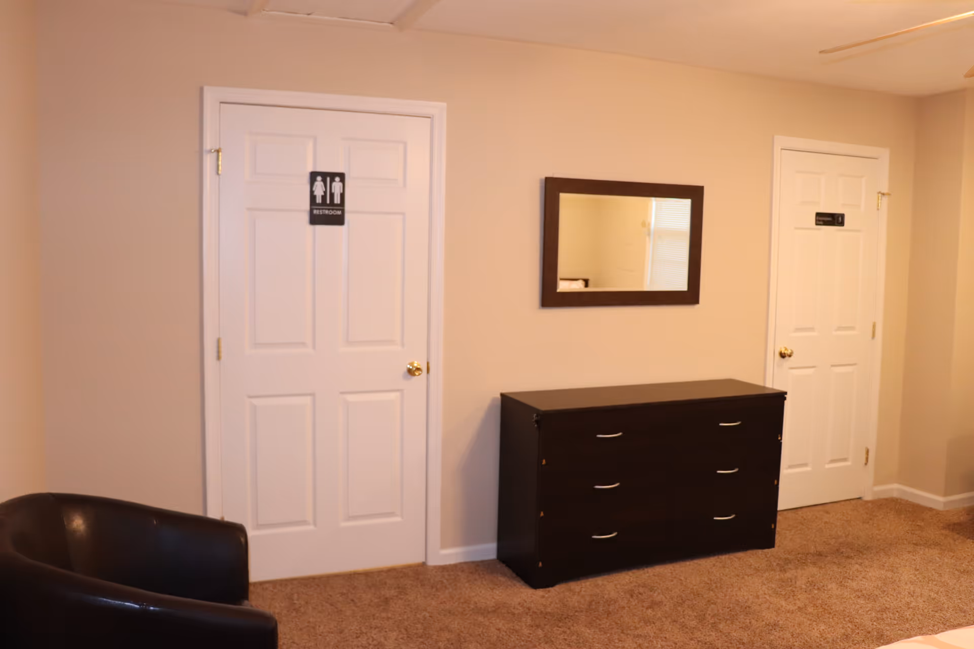 Interior view of a room with beige walls and carpeted floor, featuring two white doors. The left door has a restroom sign, and the right door has an employee only sign. Between the doors is a dark wooden dresser with six drawers and a rectangular mirror mounted above it. A dark brown leather chair is partially visible in the lower left corner.