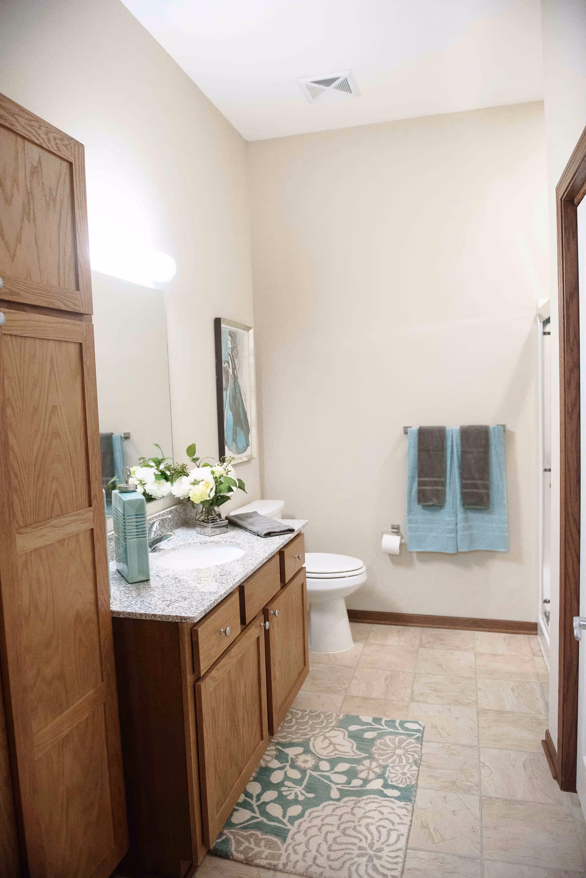 A clean and well-lit bathroom featuring a wooden vanity with a granite countertop, a sink, a large mirror, and decorative flowers. There is a toilet next to the vanity, a towel rack with two blue and two gray towels, a patterned rug on the tiled floor, and a shower stall visible on the right side.