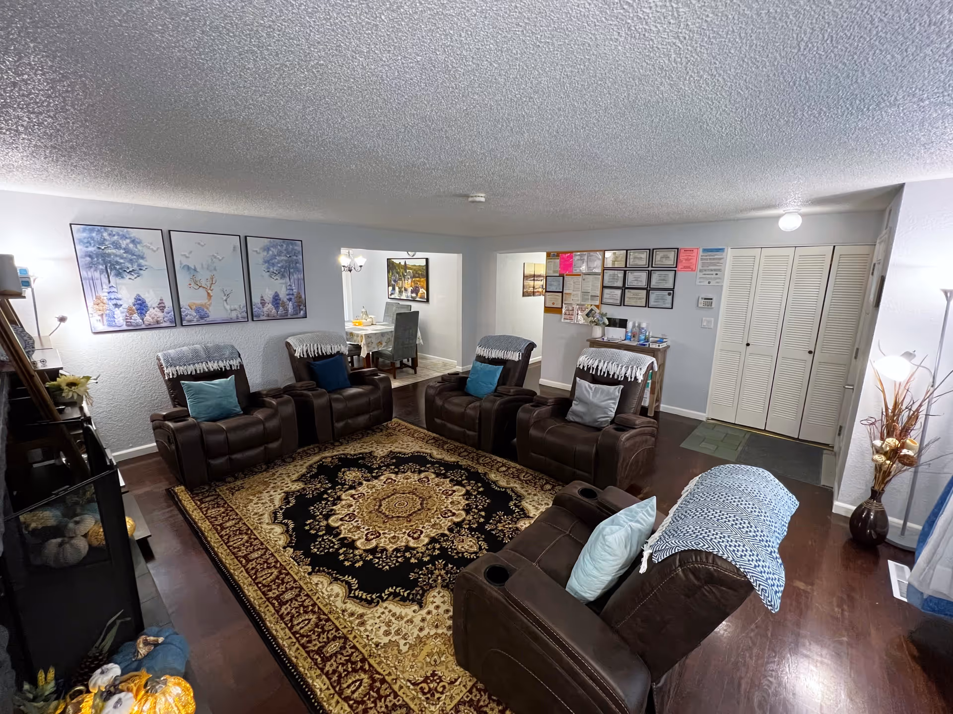 A cozy living room with six brown recliner chairs arranged in a circle on a large ornate area rug. The walls are light gray with three framed pictures of deer and trees on one side. There is a small table with certificates and notices on the wall behind it. A dining area with a table and chairs is visible through an open doorway. The room has hardwood floors and is well-lit with lamps and ceiling lights.