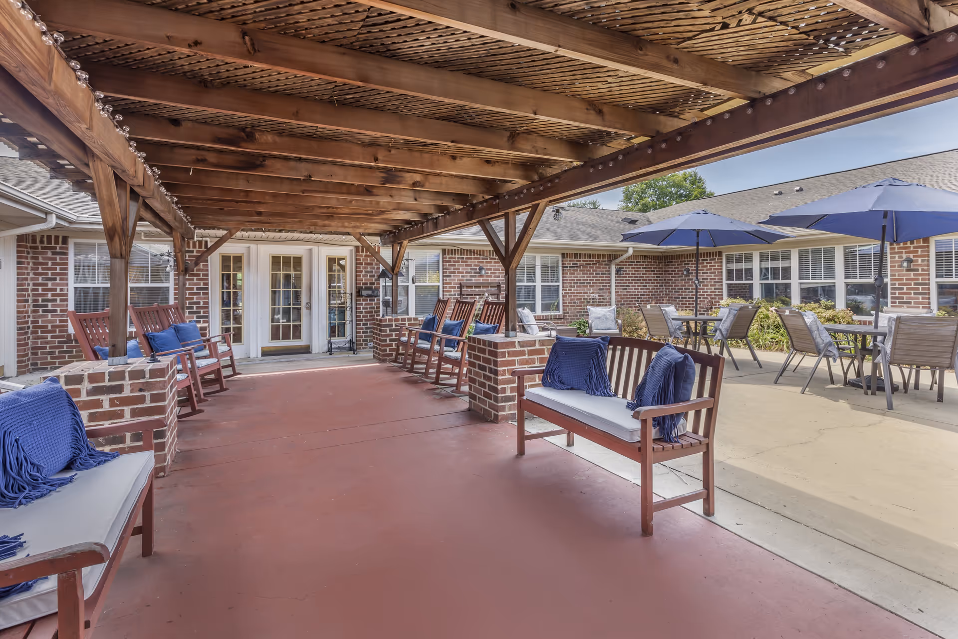 Covered outdoor patio courtyard with a wooden pergola, benches with blue cushions and umbrella-shaded tables in front of a brick building.