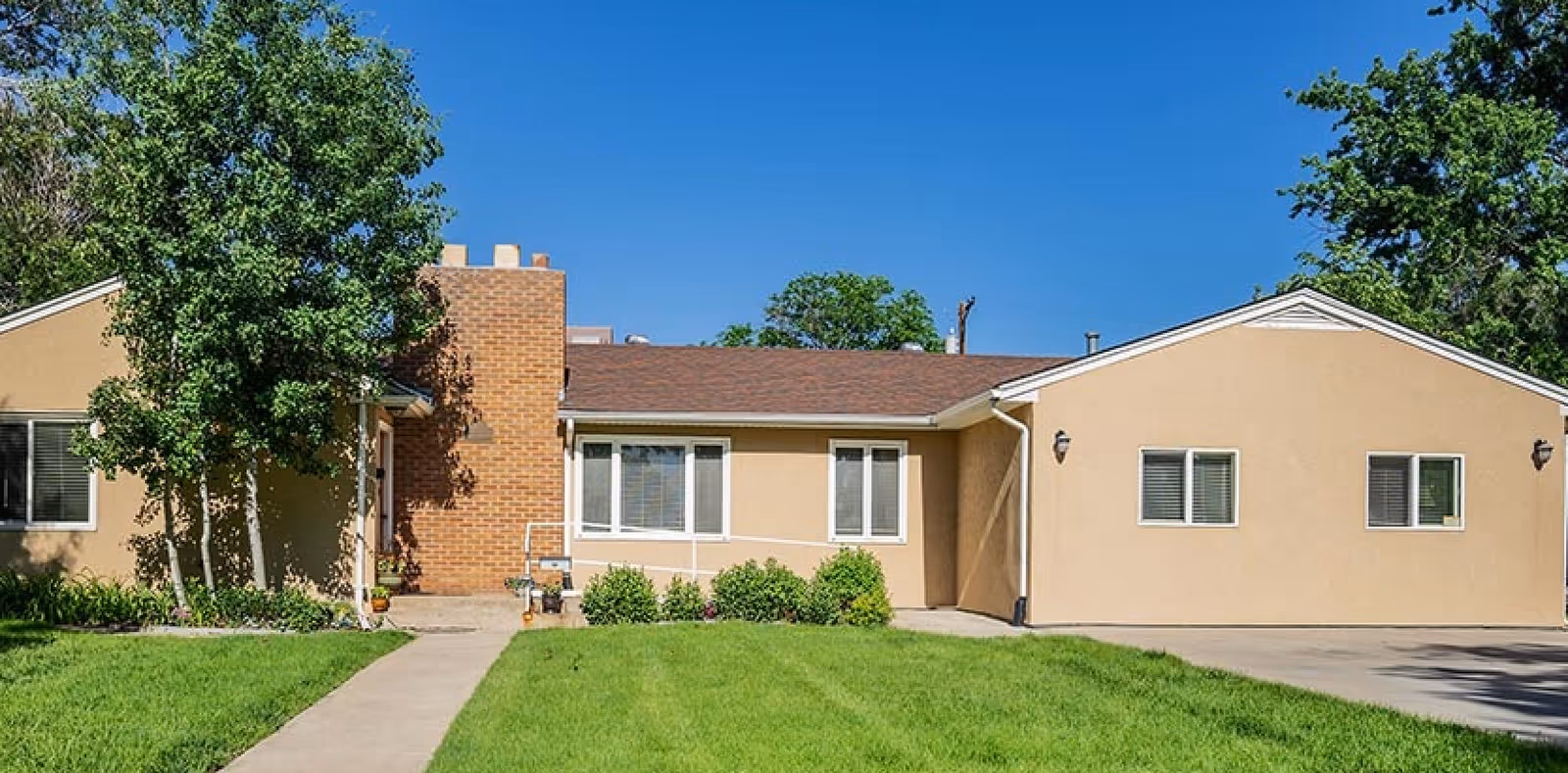 Single-story beige building with a brown shingled roof, several windows, a brick chimney, and a concrete walkway leading to the entrance. The building is surrounded by green grass, bushes, and trees under a clear blue sky.