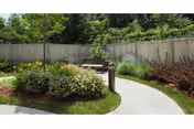 A peaceful outdoor garden area with a curved concrete pathway, surrounded by green shrubs, flowering plants, and trees. There is a wooden bench along the path and a tall light post nearby. A wooden fence encloses the garden space.