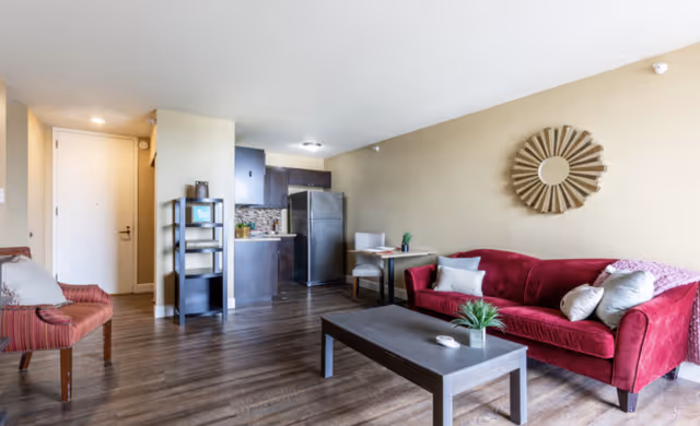 A cozy living area with a red sofa adorned with pillows and a throw blanket, a dark wooden coffee table with a small plant and decorative items, a small dining table with two chairs, and a compact kitchen with dark cabinets and a stainless steel refrigerator. The room has wooden flooring and beige walls, with a decorative round mirror hanging above the sofa.