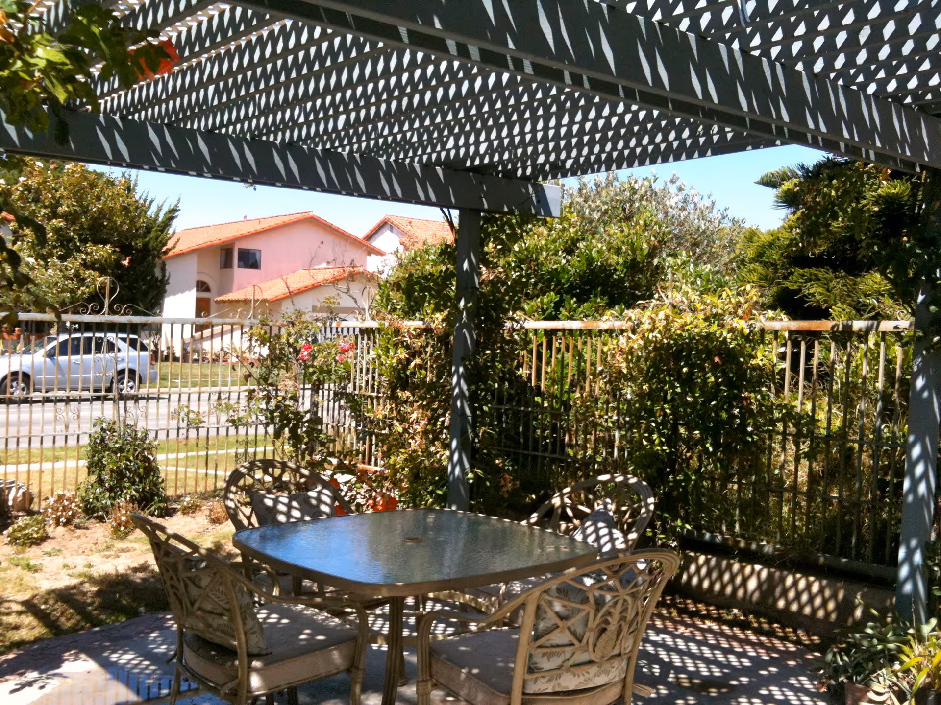 Outdoor patio area with a glass-top table and four cushioned chairs under a pergola casting patterned shadows. Surrounding the patio are green bushes and a metal fence, with houses and a parked car visible in the background under a clear blue sky.