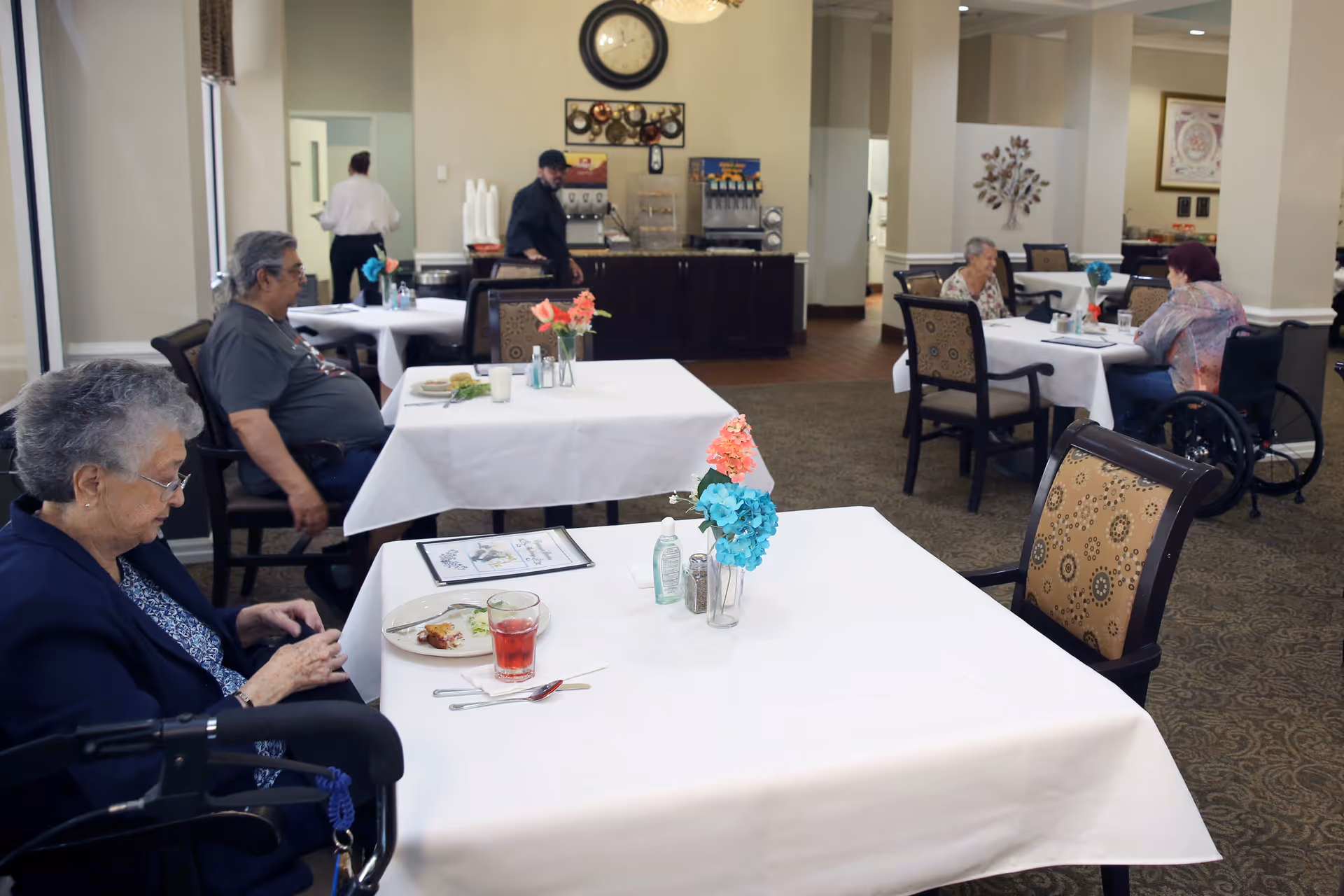 A dining room in a senior living facility with several elderly residents seated at tables covered with white tablecloths. There are flower vases on the tables, and a beverage station with a clock on the wall in the background. Two staff members are visible, one standing near the beverage station and another walking away.