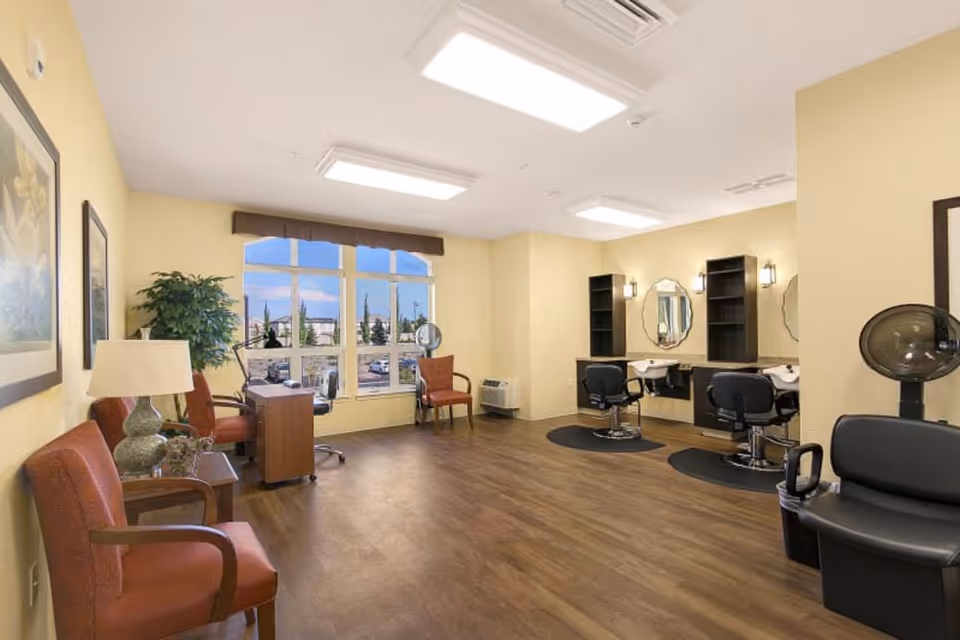 A spacious salon area in a senior living facility with two styling stations featuring mirrors, chairs, and sinks. There are additional seating chairs along the left wall, a desk near large windows letting in natural light, and a hair dryer chair on the right side. The room has wooden flooring and soft yellow walls.