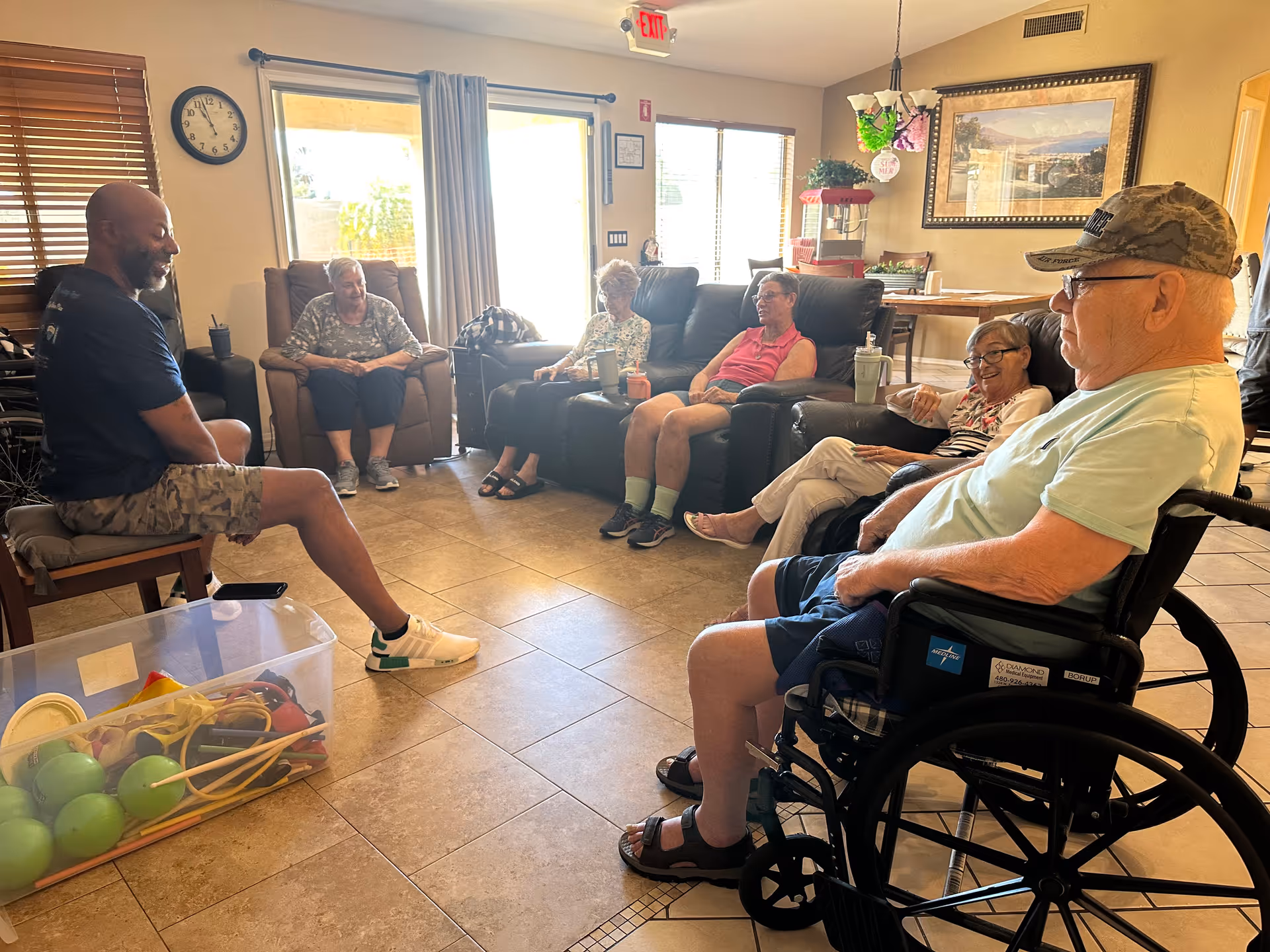 A group of six elderly people and one younger man sitting in a living room area of an assisted living facility. The elderly individuals are seated on recliners and a wheelchair, engaging in conversation. The room has tiled floors, large windows with blinds, a clock on the wall, and a popcorn machine in the background.