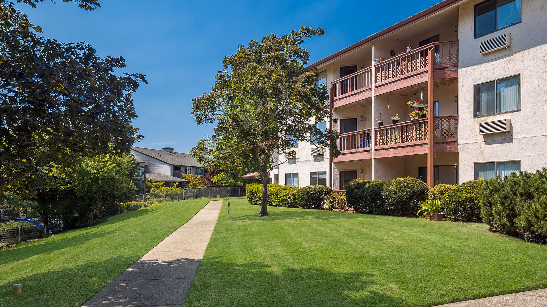 Exterior view of a multi-story apartment building with balconies facing a manicured lawn and a concrete walkway.