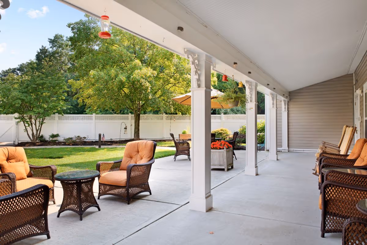 Covered patio with wicker chairs and orange cushions overlooking a landscaped yard with trees and a white fence.