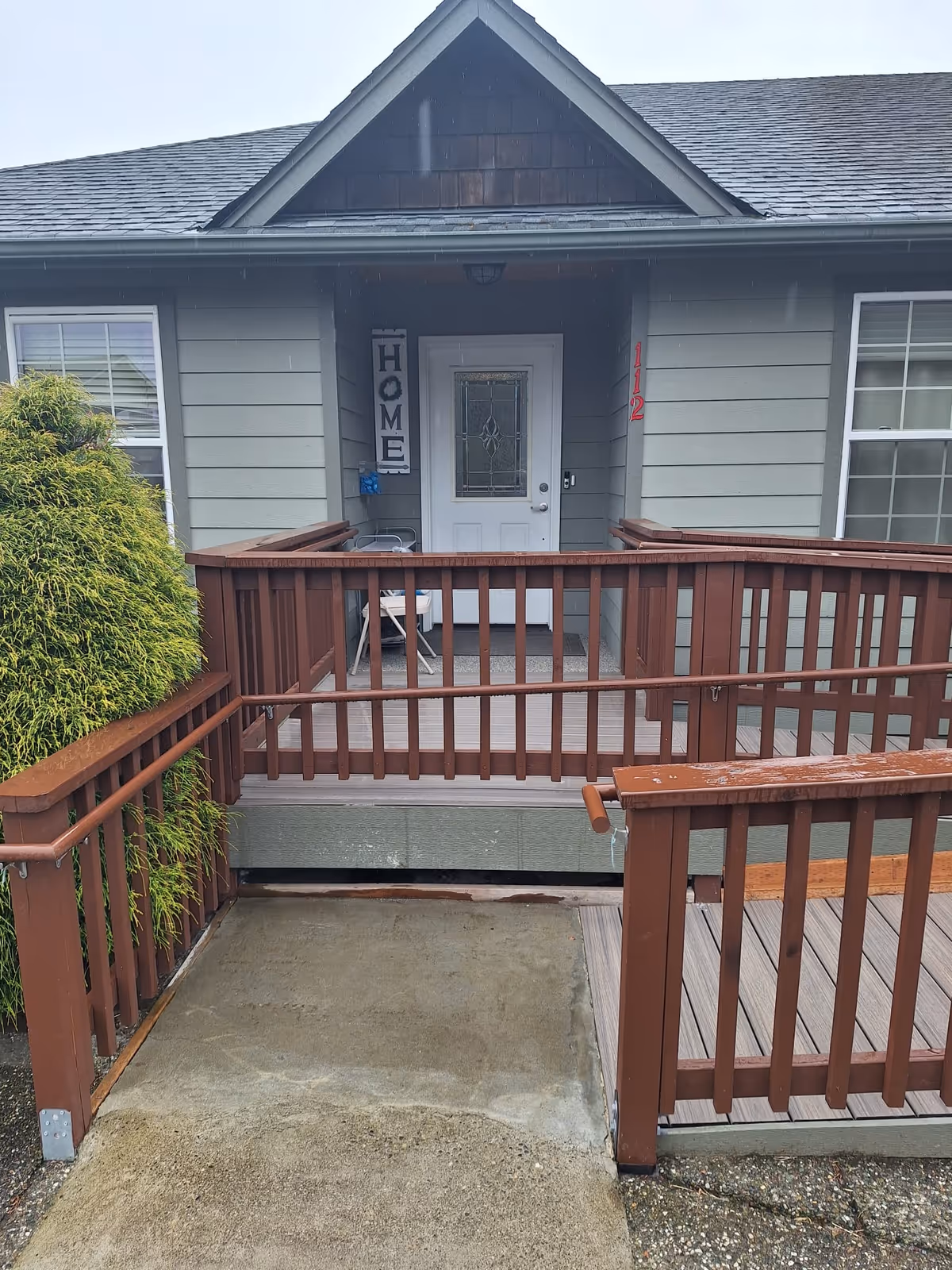 Front entrance of a single-story house with a wooden wheelchair ramp and railings leading to a white door displaying a 'HOME' sign.