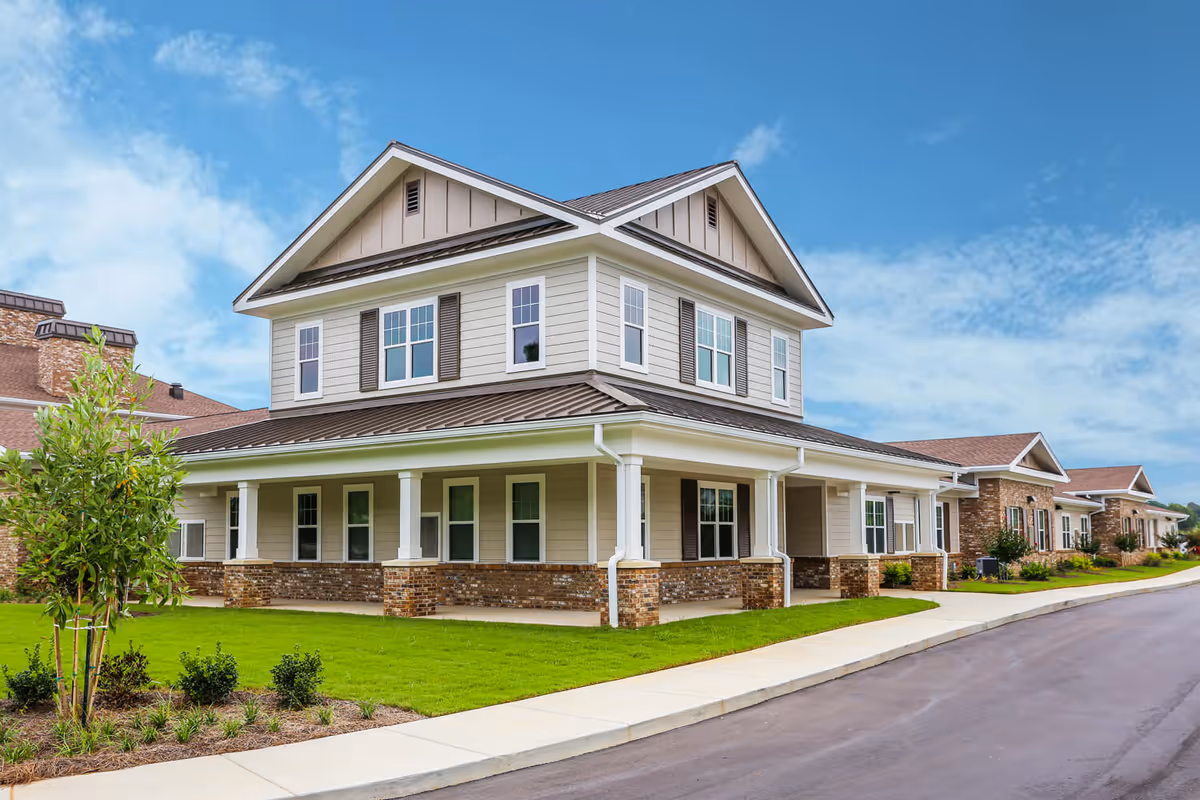 Exterior view of a two-story senior living facility building with beige siding, brown roof, and white trim. The building has multiple windows and a covered porch supported by white columns. There is a well-maintained green lawn and a paved driveway in front under a partly cloudy blue sky.