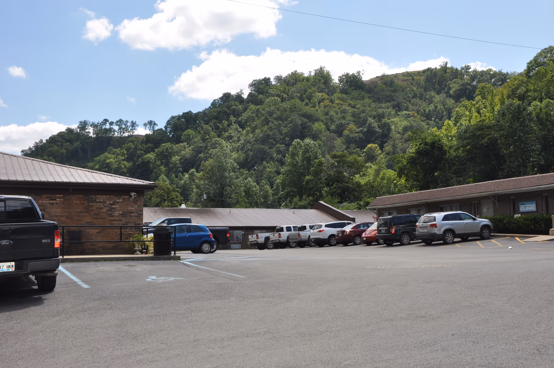Parking lot with cars in front of a low brick healthcare building and a wooded hillside in the background.