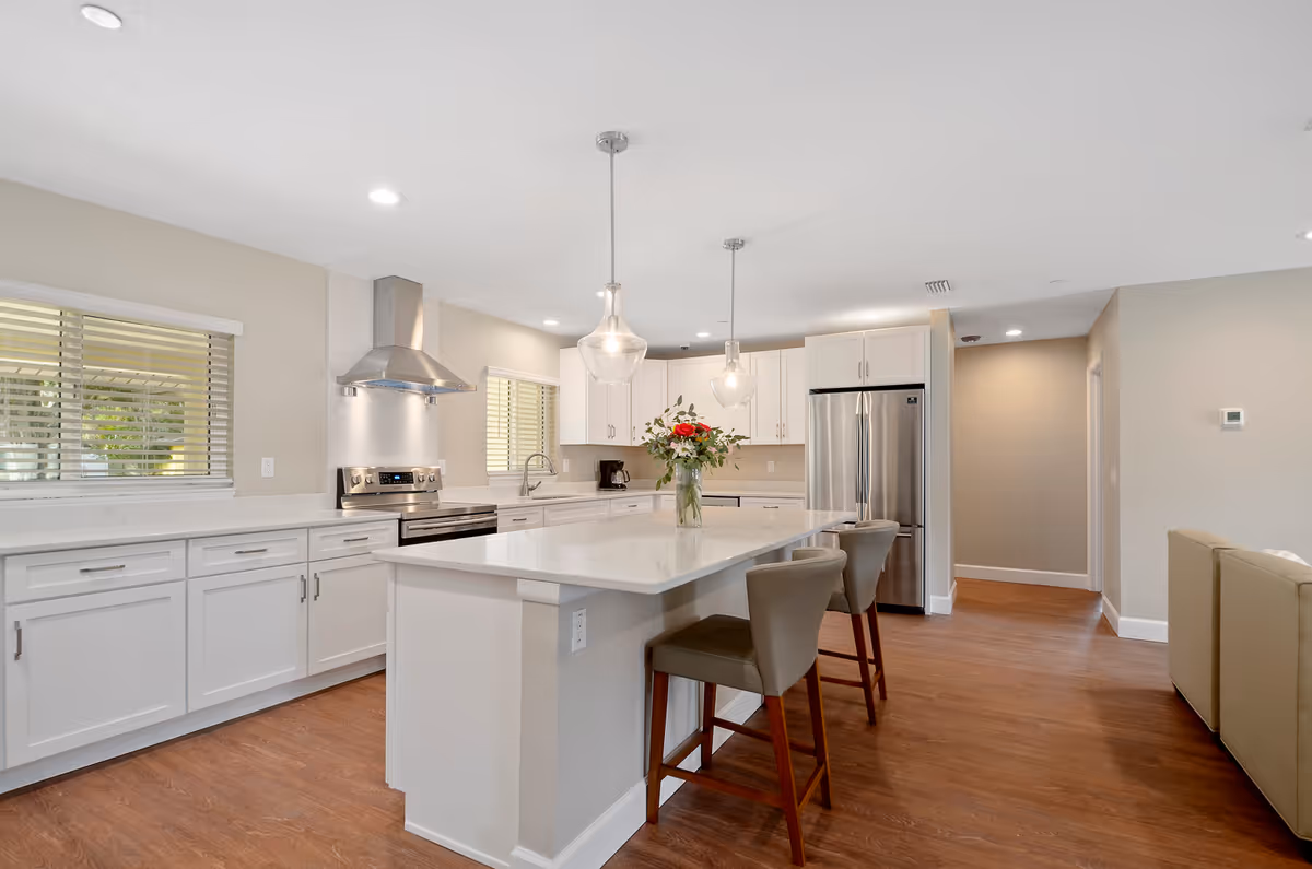 Bright modern open kitchen with white cabinets, a large island with stools and pendant lights, stainless steel appliances, and wood floors.