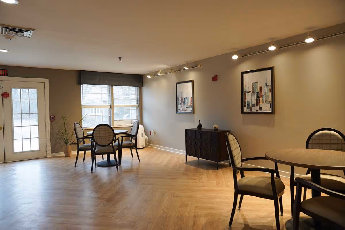 A well-lit interior room with wooden flooring featuring two round tables each surrounded by four cushioned chairs. There are two framed abstract paintings on the beige walls, a small dark wooden cabinet with decorative items, a large window with blinds, and a glass door leading outside. Ceiling lights illuminate the space.
